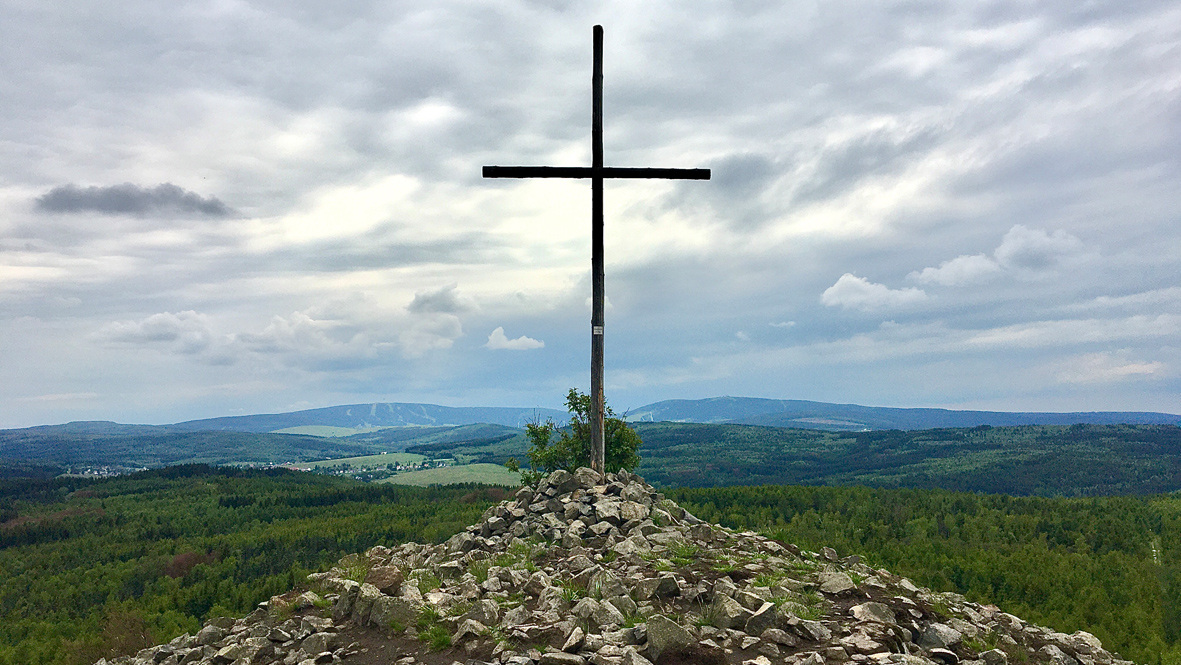 Das Gipfelkreuz auf dem Velký Špičák (Großer Spitzberg). Dahinter rechts der 1.215 Meter hohe Fichtelberg in Deutschland und links der 1.244 Meter hohe Klínovec (Keilberg) in Tschechien.